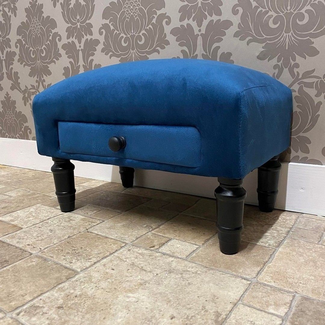 Blue velvet footstool with a curved design, featuring a small drawer and black wooden legs, measuring 25 x 45 x 28 cm, set against a patterned wall.