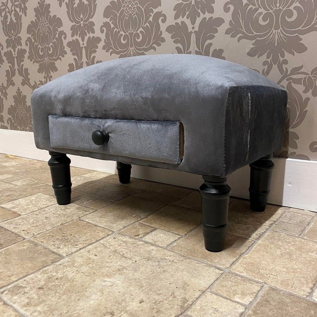 Grey velvet footstool with a curved top, featuring a small drawer and black wooden legs, measuring 25 x 45 x 28 cm, set against a patterned wallpaper background.
