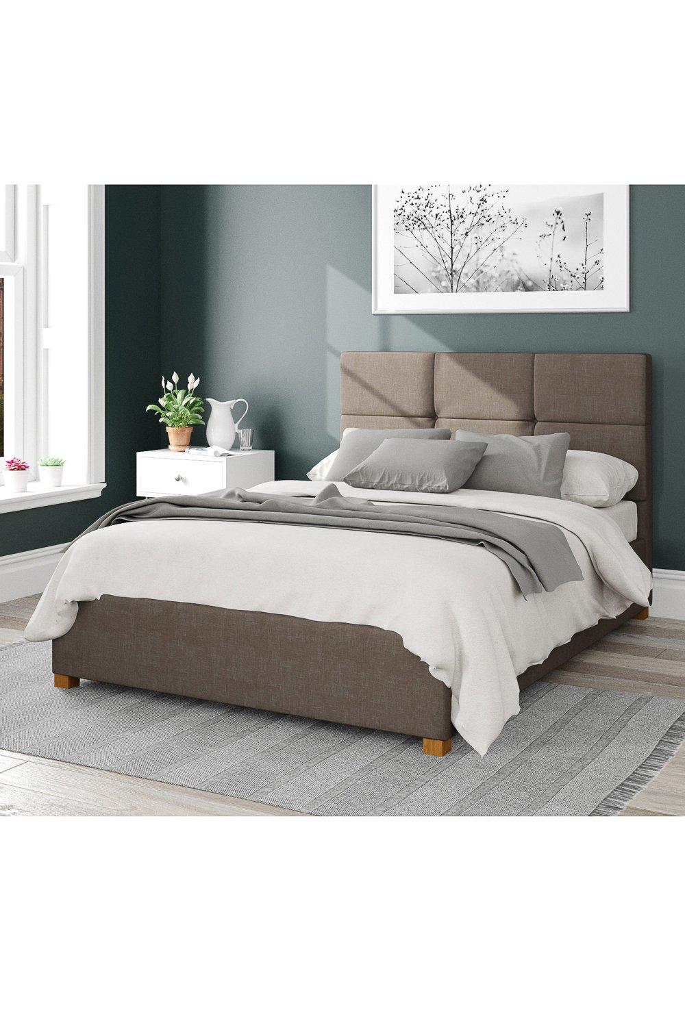 Caine Upholstered Ottoman Storage Bed in Malham weave fabric features a soft brown exterior, a padded headboard, and wooden legs, styled with neutral bedding and a cozy atmosphere.