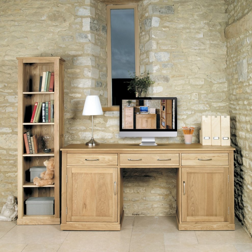 Large twin pedestal desk in brown oak with two drawers and a spacious surface, accompanied by a wooden bookshelf, lamp, and computer monitor in a stone-walled room.