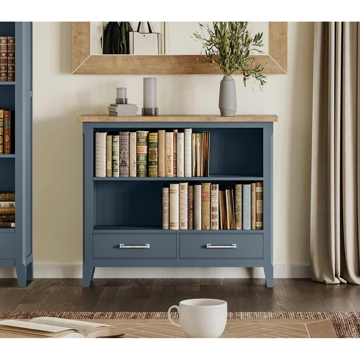 Low bookcase in cobalt blue with a natural wood top, featuring two open shelves for books and two drawers with silver handles, set against a light wall and decorative elements.