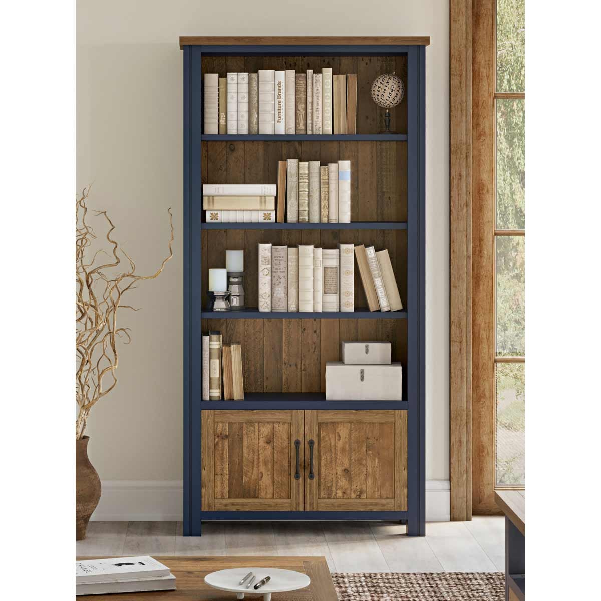 Large open bookcase with blue frame and natural wood doors, featuring five shelves filled with books, and a decorative sphere on top, set against a bright room backdrop.