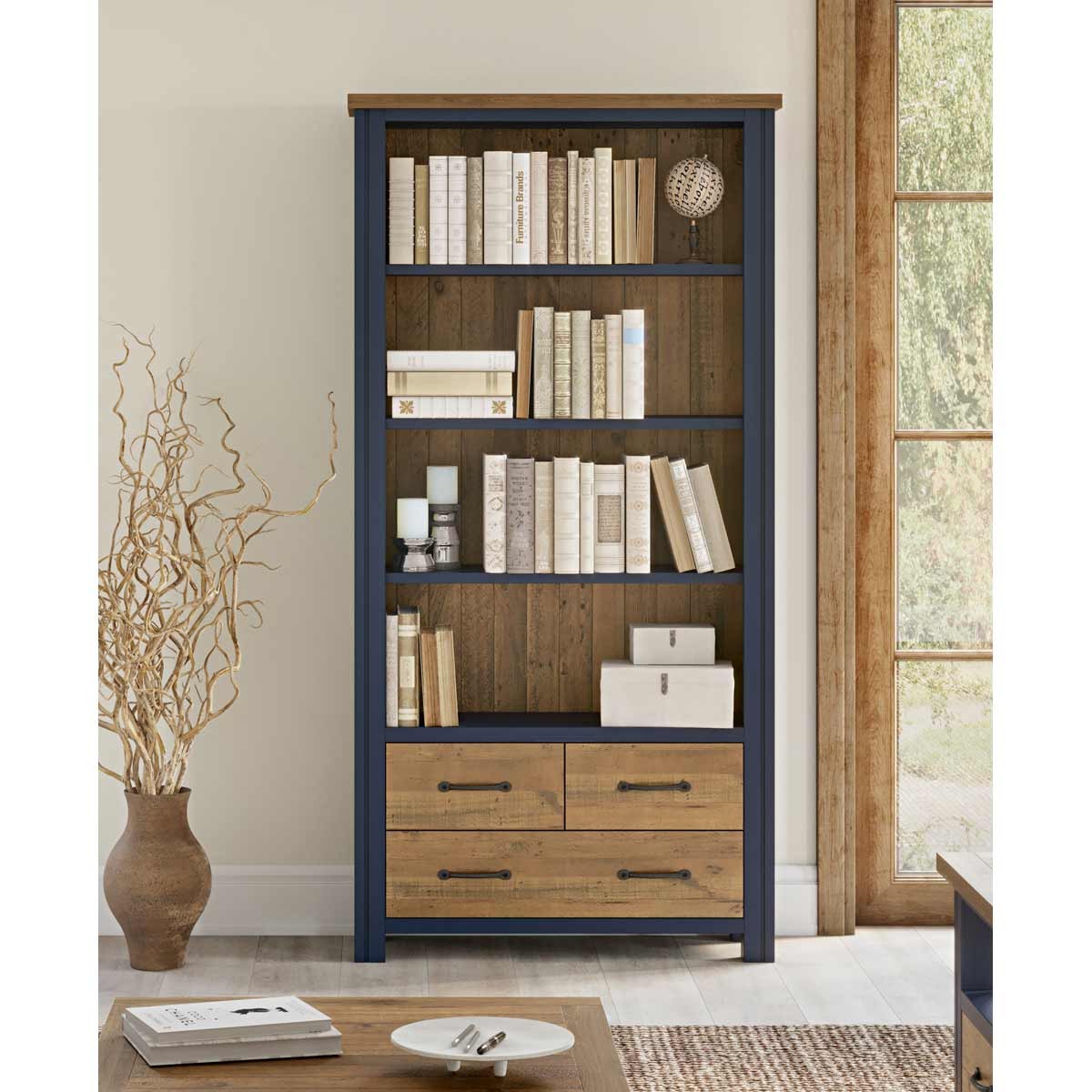 Large open bookcase in blue with a wooden top, featuring multiple shelves filled with books and two lower drawers with metal handles, set against a bright room backdrop.