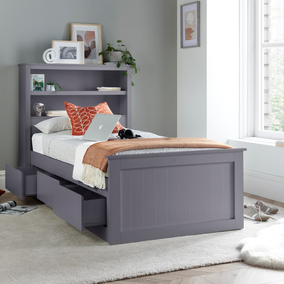 Grey wooden single bed with three drawers and a bookcase headboard, featuring a cozy white bedding set, decorative pillows, and a laptop on top, set against a light wall.