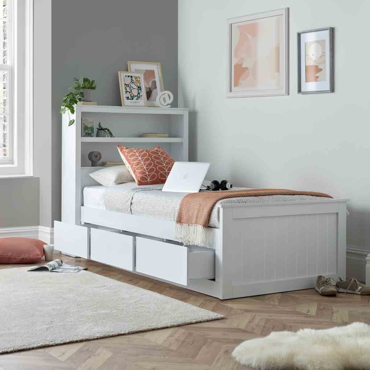 White wooden bed with three drawers, featuring a pocket mattress, soft bedding, and decorative pillows, set against a light green wall in a cozy room.