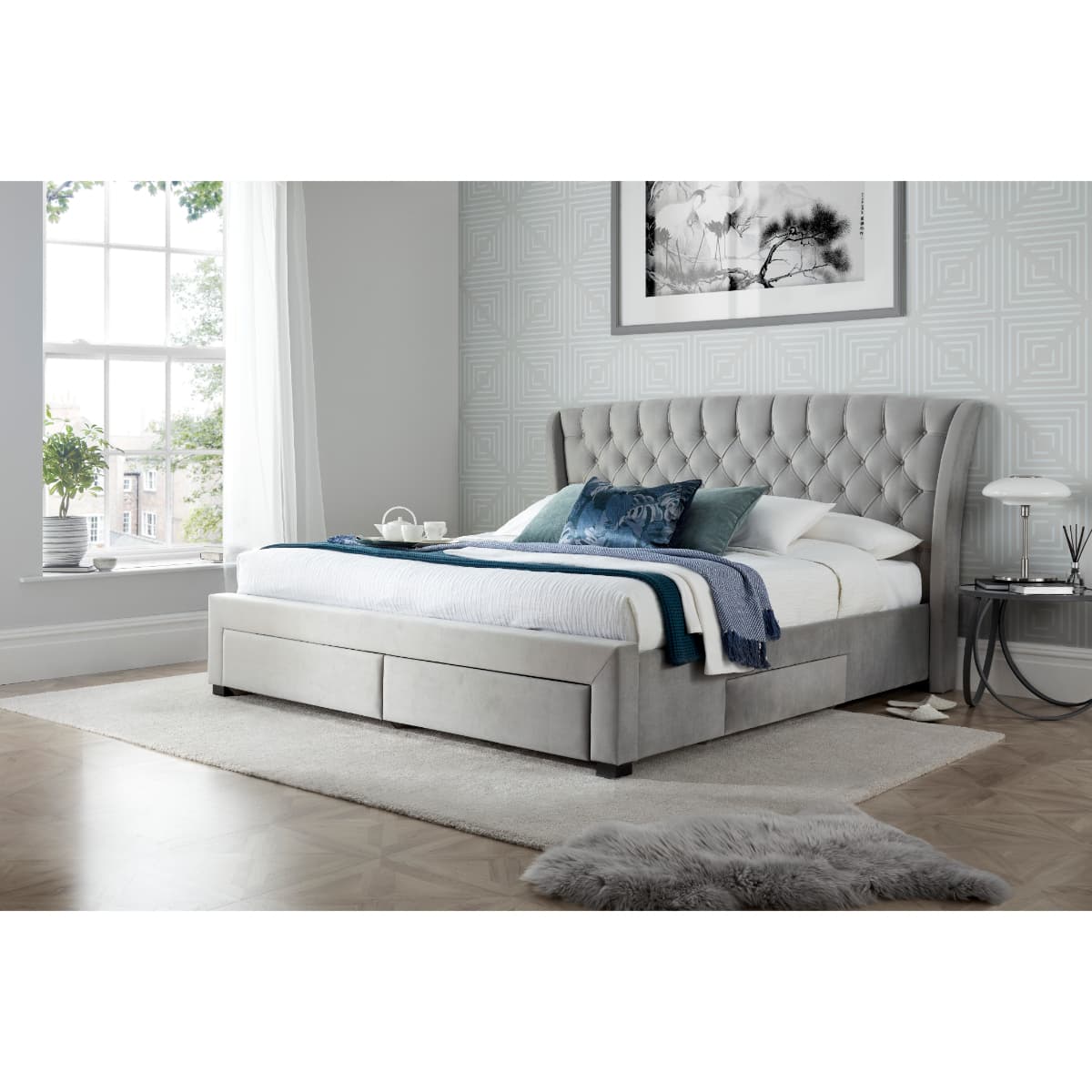 Elegant grey velvet storage bed with a tufted headboard, featuring four spacious drawers for storage, set in a bright room with large windows and modern decor.