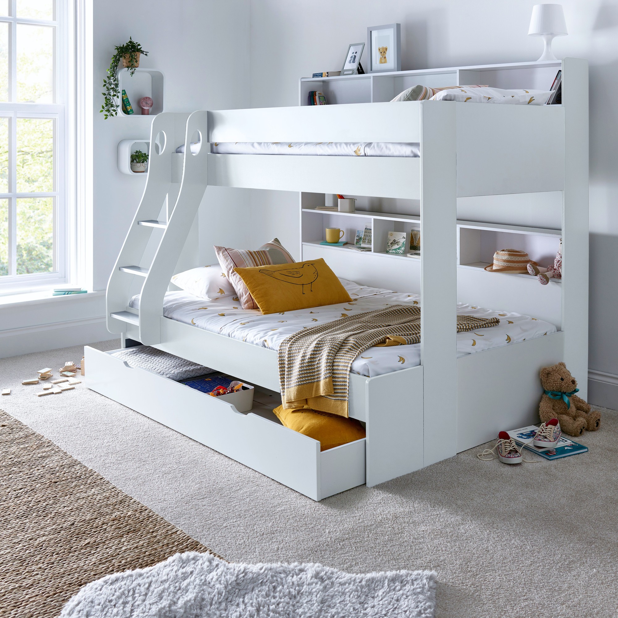 White triple sleeper bunk bed with orthopaedic mattresses, featuring a ladder, open shelving, and a storage drawer, set in a bright room with a cozy rug and decorative cushions.
