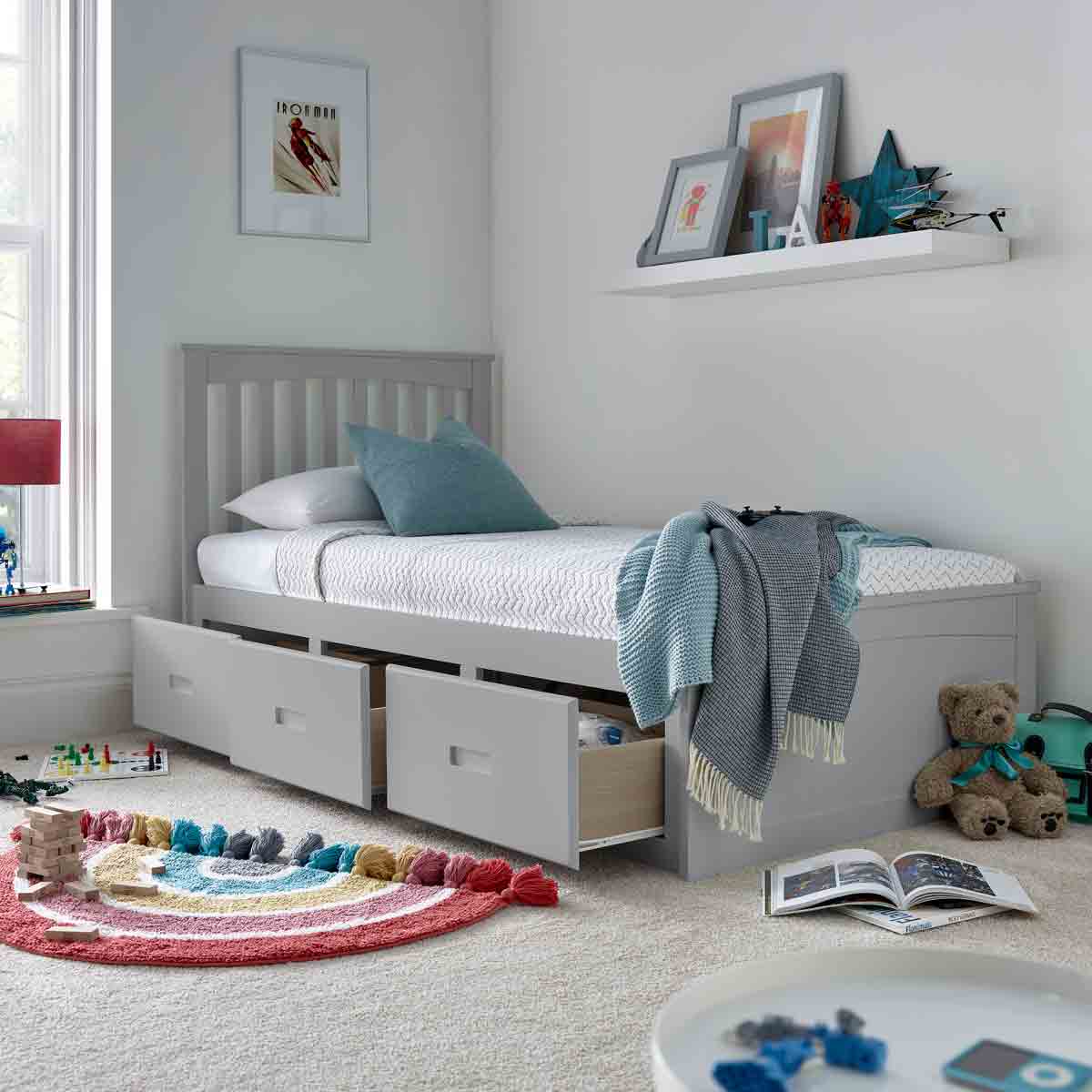 Grey wooden bed with three drawers underneath, featuring a slatted headboard, a cozy mattress, and a light blue throw blanket, set in a bright, modern room.
