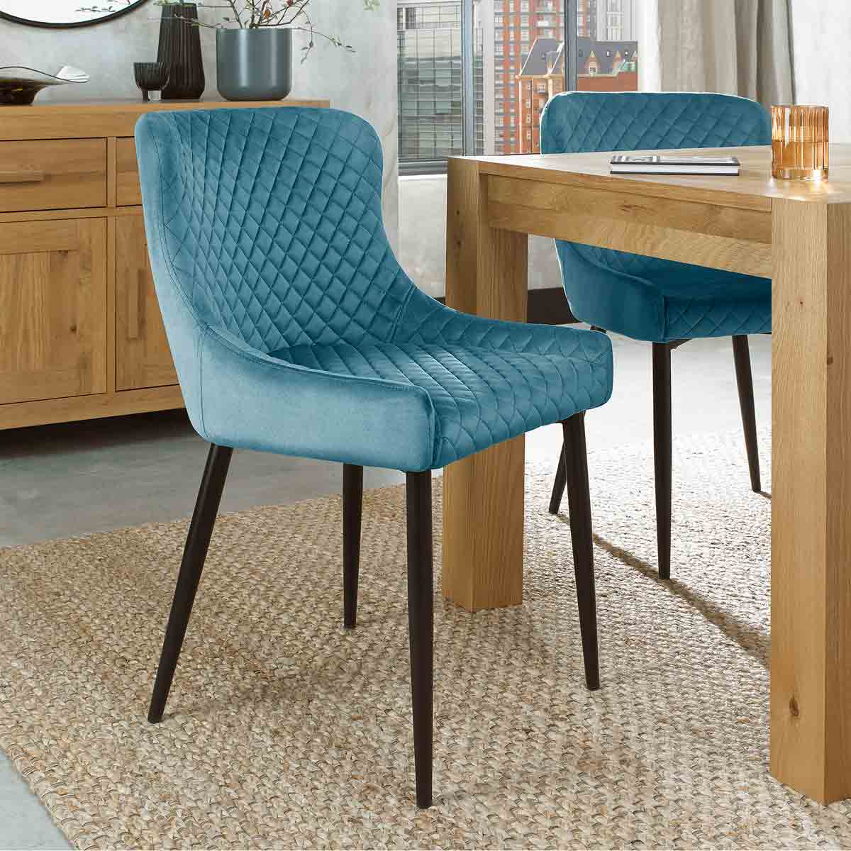 Petrol blue velvet fabric chair with a diamond quilted pattern and sleek black legs, positioned next to a wooden table on a textured rug.
