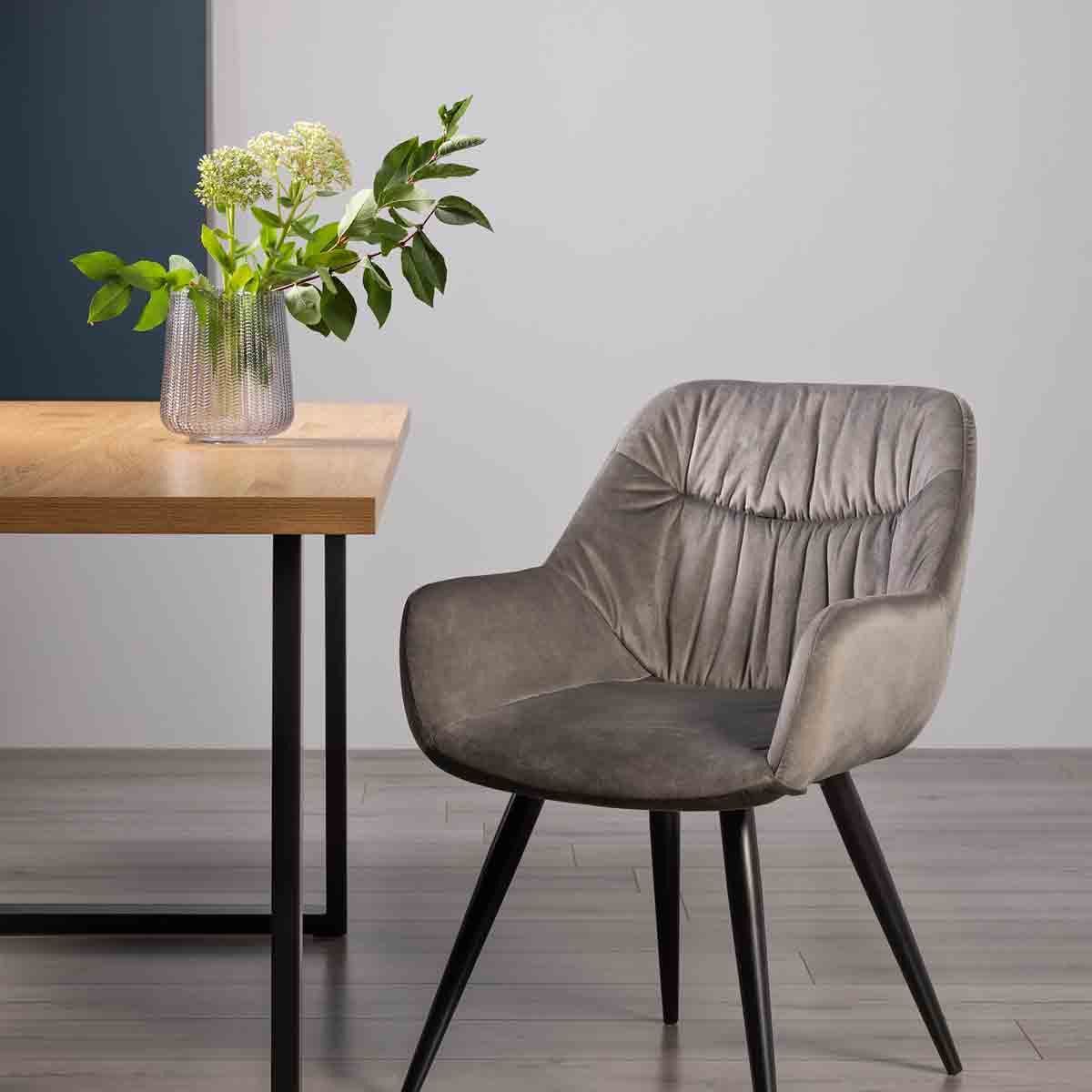 Grey velvet fabric chair with black legs, featuring a tufted design, positioned beside a wooden table with a vase of greenery.