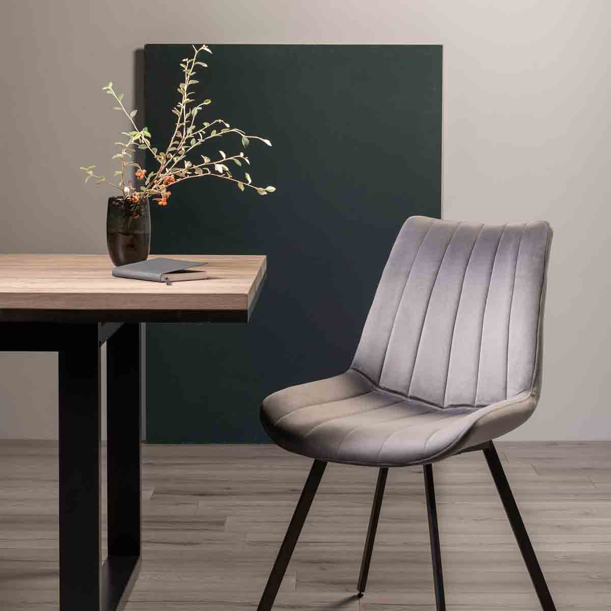 Pair of grey velvet chairs with black legs, featuring vertical stitching, positioned beside a wooden table with a vase of flowers and a notebook against a neutral backdrop.