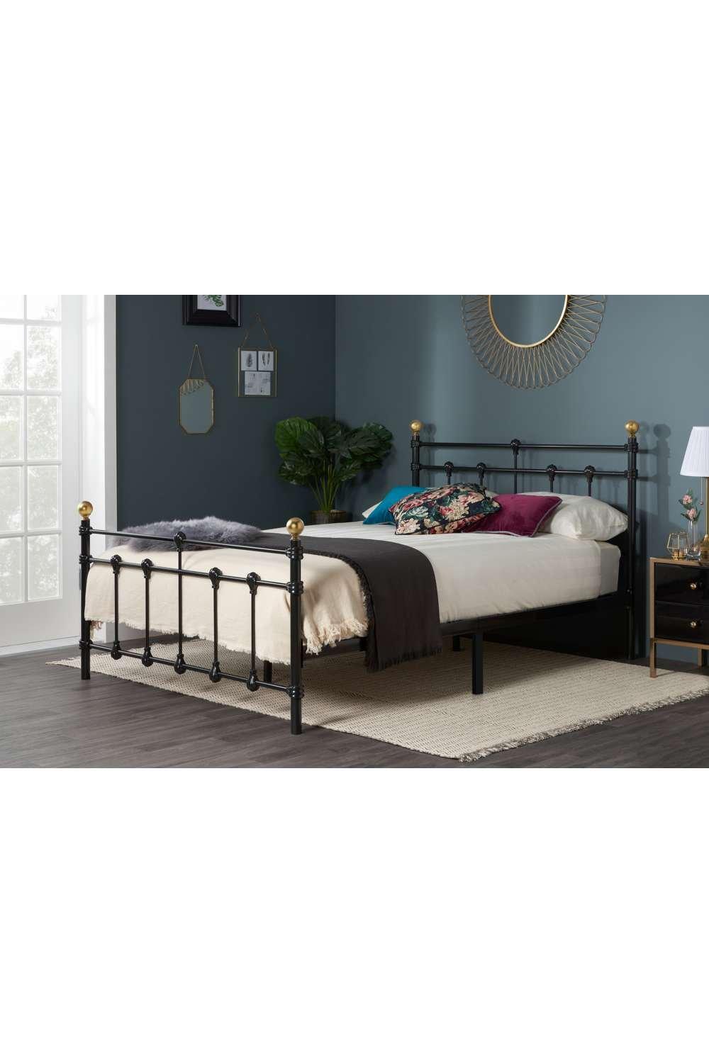 Black metal bed frame with gold finials, featuring a classic design, sturdy slatted base, and a stylish headboard, set against a dark blue wall and light-colored rug.