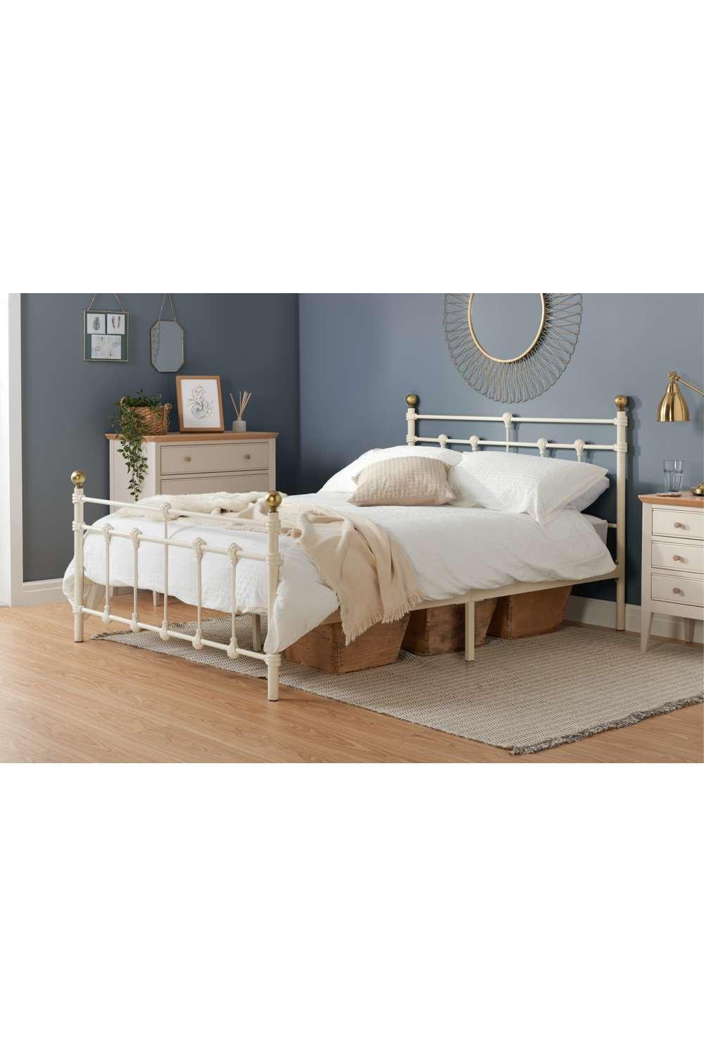 Cream-colored Birlea Atlas bed with a small double frame, featuring decorative brass finials, a metal headboard, and a soft, neutral bedding set against a blue wall.