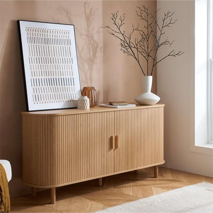 Brown fluted sideboard with two doors and wooden legs, complemented by a white vase and a framed artwork against a beige wall, enhancing a modern interior aesthetic.