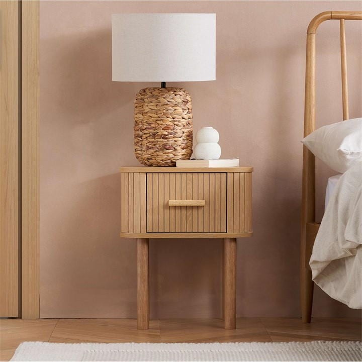 Oak brown wooden bedside table with fluted design, featuring a single drawer and round wooden handle, topped with a woven lamp and decorative white stones.