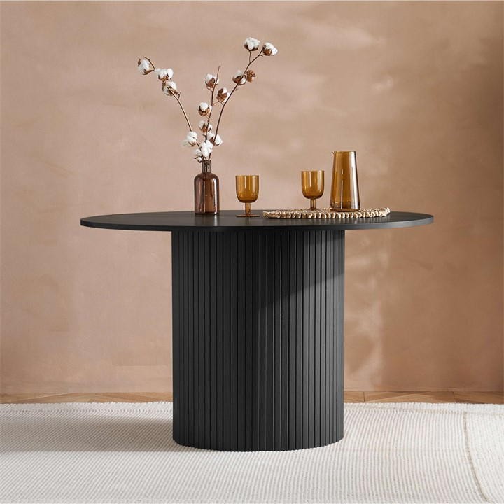 Round black fluted dining table with a cylindrical base, styled with decorative glassware and a vase of cotton branches, set against a warm, neutral background.