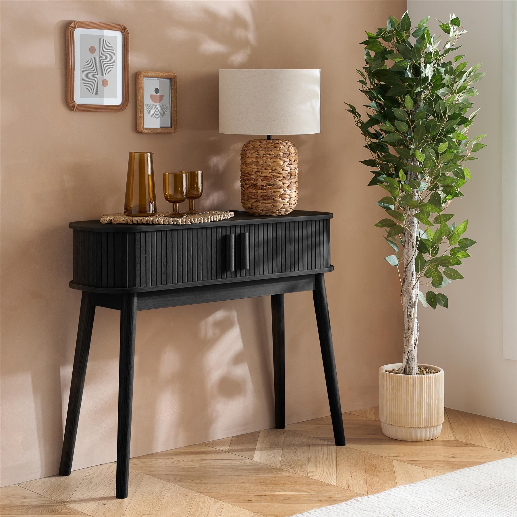 Fluted black console table with two doors, set against a beige wall, accompanied by a lamp, decorative vases, and a potted plant.