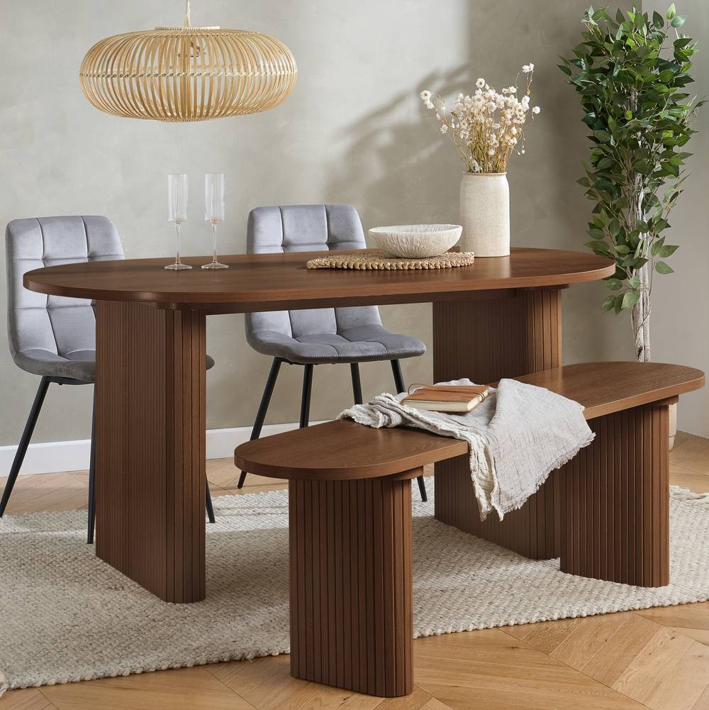 Brown oval fluted dining table with two grey upholstered chairs and a matching bench, set on a light wooden floor with a decorative plant and pendant light above.