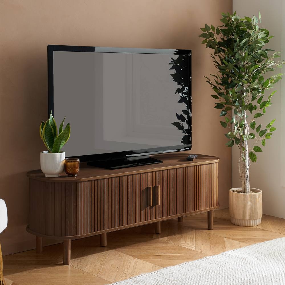 Birlea Axel Fluted TV Unit in walnut brown features a sleek, curved design with fluted detailing, two doors, and stands on tapered legs, complemented by a potted plant nearby.