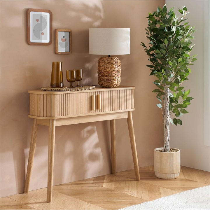 Brown fluted console table with two drawers, wooden legs, styled with a lamp, decorative vases, and a potted plant against a light-colored wall.