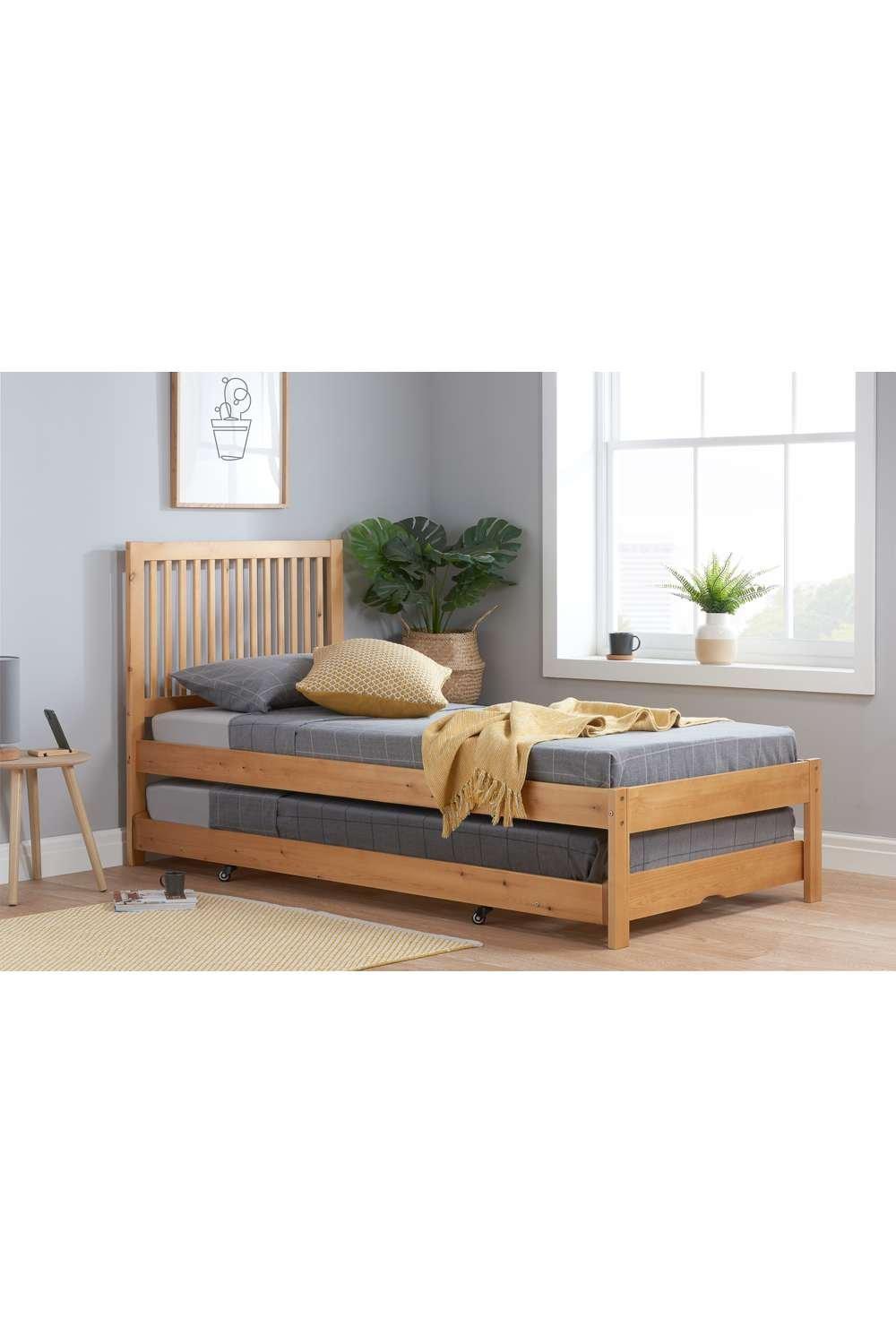 Light wood trundle bed with slatted headboard, featuring a grey mattress and a cozy yellow throw, set in a bright room with plants and natural light.