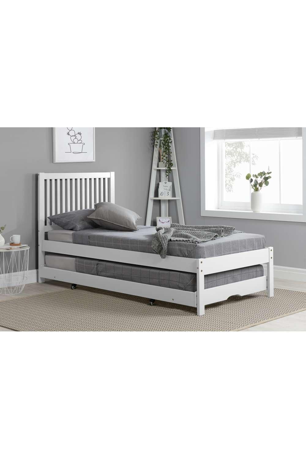 White wooden bed frame with a slatted headboard and a pull-out trundle underneath, featuring gray bedding and set against a light gray wall with a decorative plant.