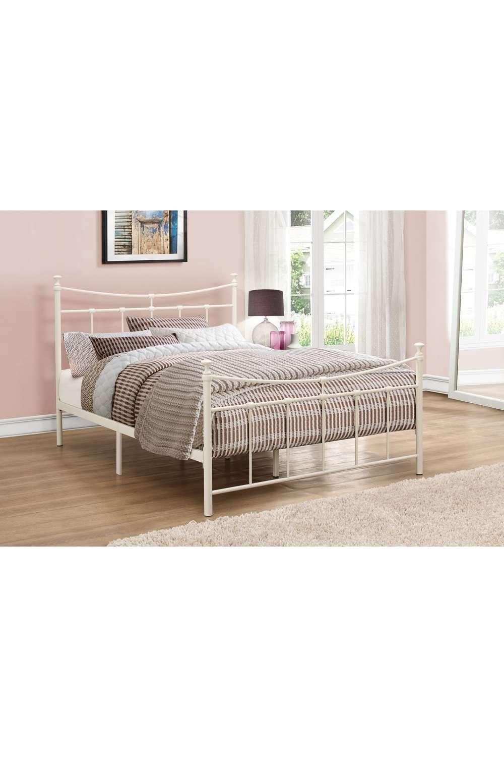Cream metal bed frame with a small double size, featuring a classic design, round finials, and a slatted headboard, set against a light pink wall and wooden floor.