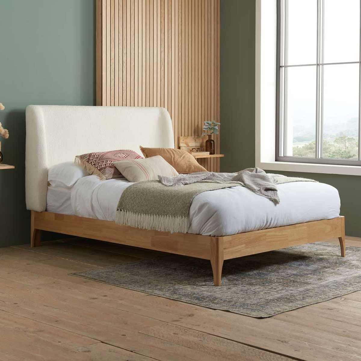 White upholstered king bed frame with a soft, curved headboard, wooden legs, and a natural wood finish, set against a green wall and a large window.