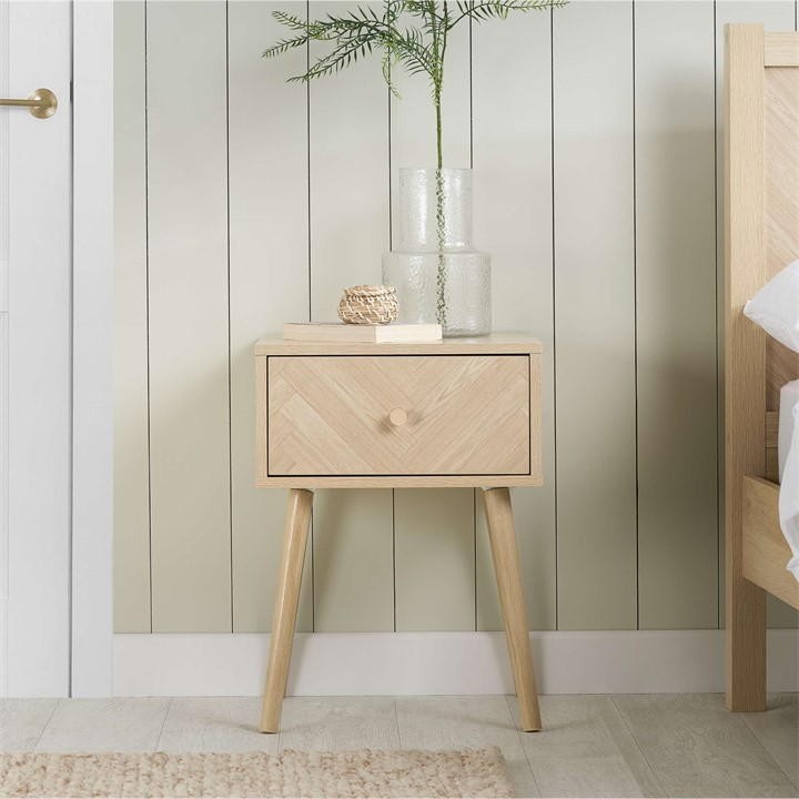 Nude wooden bedside table with a single drawer featuring a herringbone pattern, supported by tapered legs, and adorned with a plant and glass vases on top.