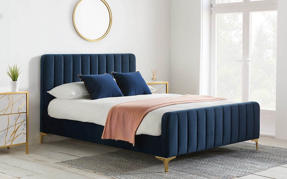 Navy blue fabric king size bed frame with vertical tufting, gold legs, and a stylish design, complemented by a white bedding set and decorative pillows.