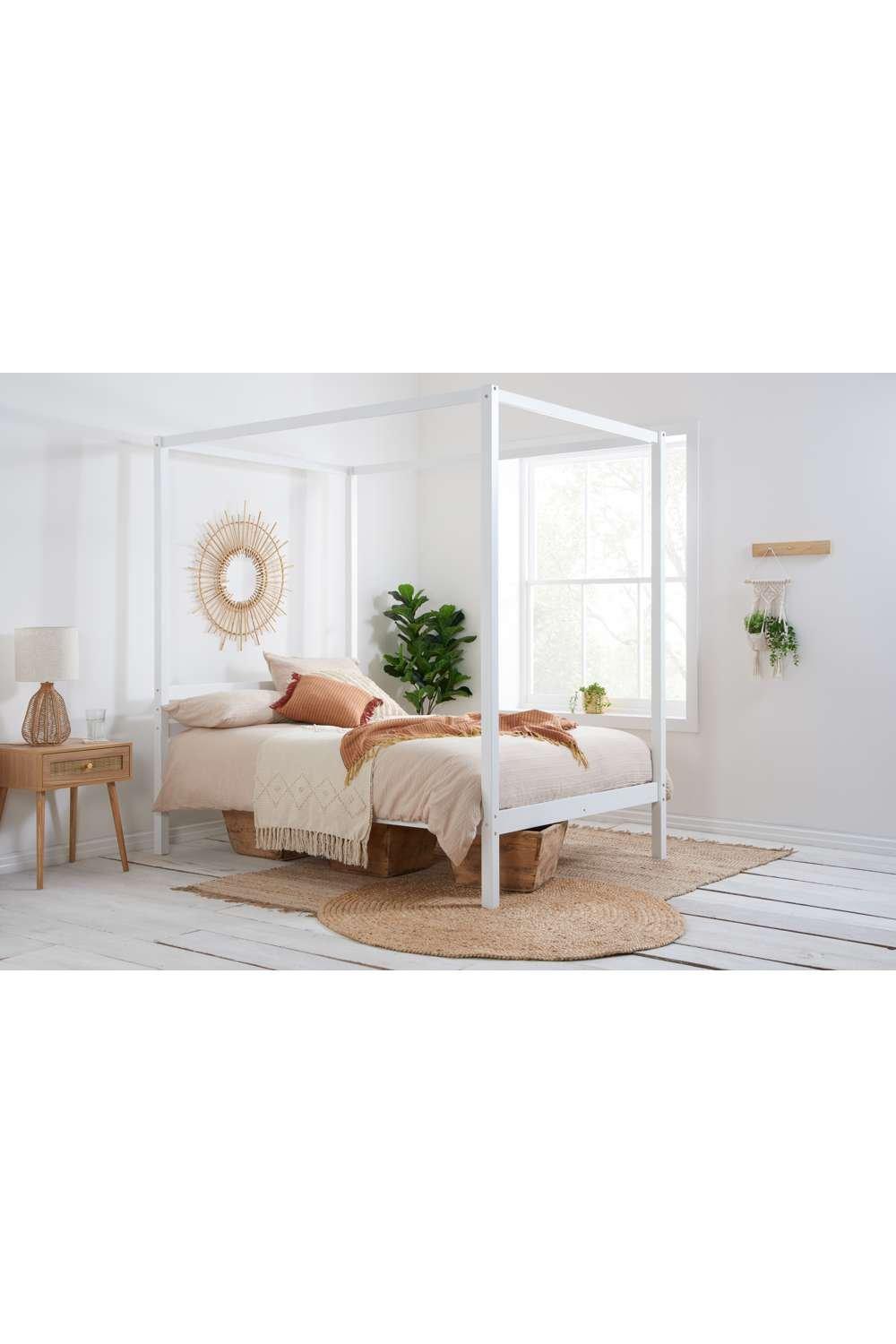 Four-poster bed frame in white solid wood, featuring a canopy design, complemented by a cozy beige bedding set and a stylish bedside lamp in a bright, airy room.