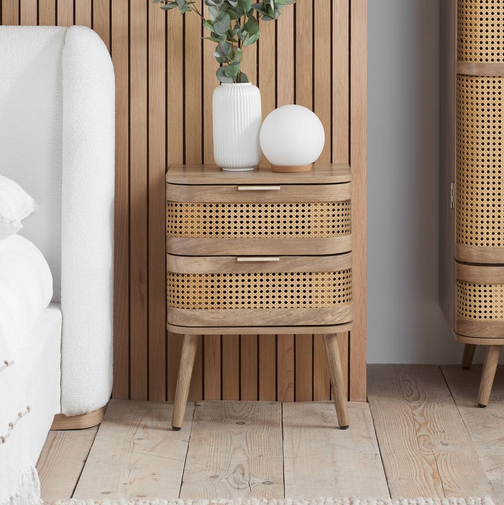Beige rattan bedside cabinet with two drawers, featuring a natural wood frame and tapered legs, complemented by a white vase and round lamp on top.