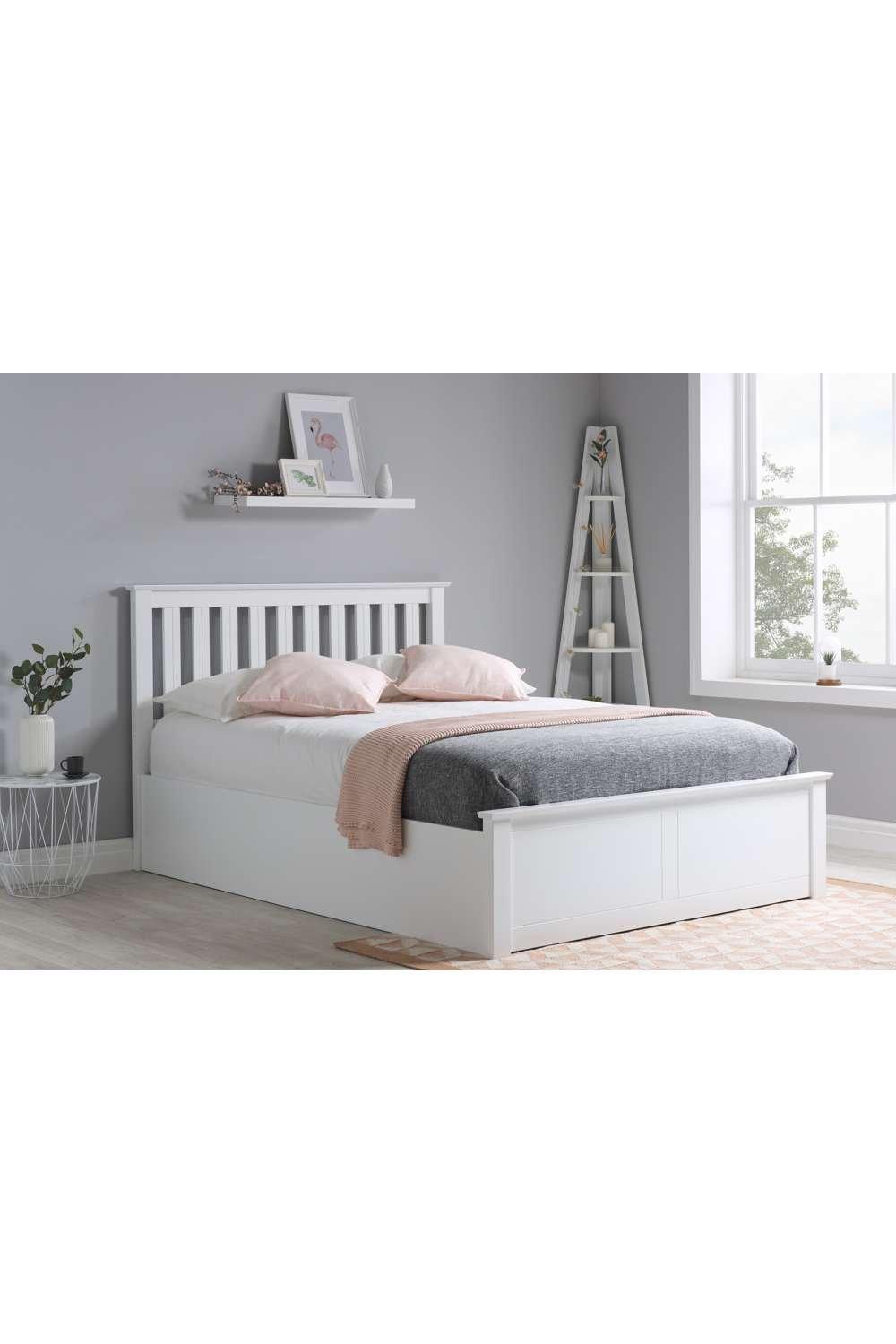 Solid white wooden ottoman storage bed frame with a gas lift mechanism, featuring a slatted headboard and a cozy gray and pink bedding arrangement.
