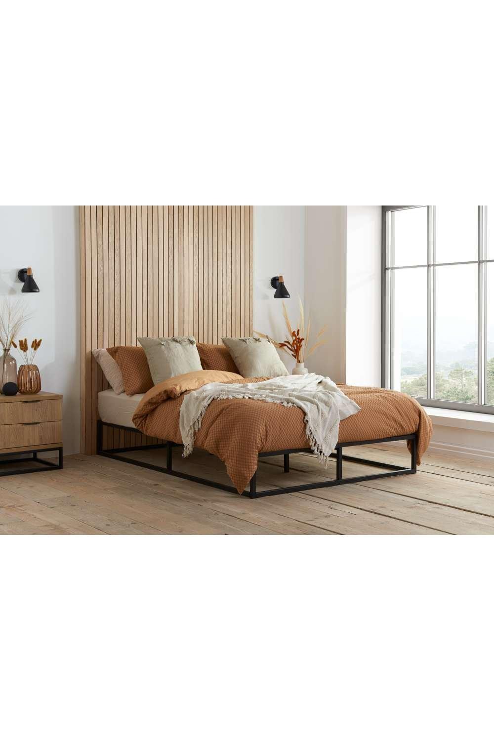 Black metal platform bed with a minimalist design, featuring a sturdy frame and a low profile, styled with warm-toned bedding and decorative pillows against a wooden accent wall.