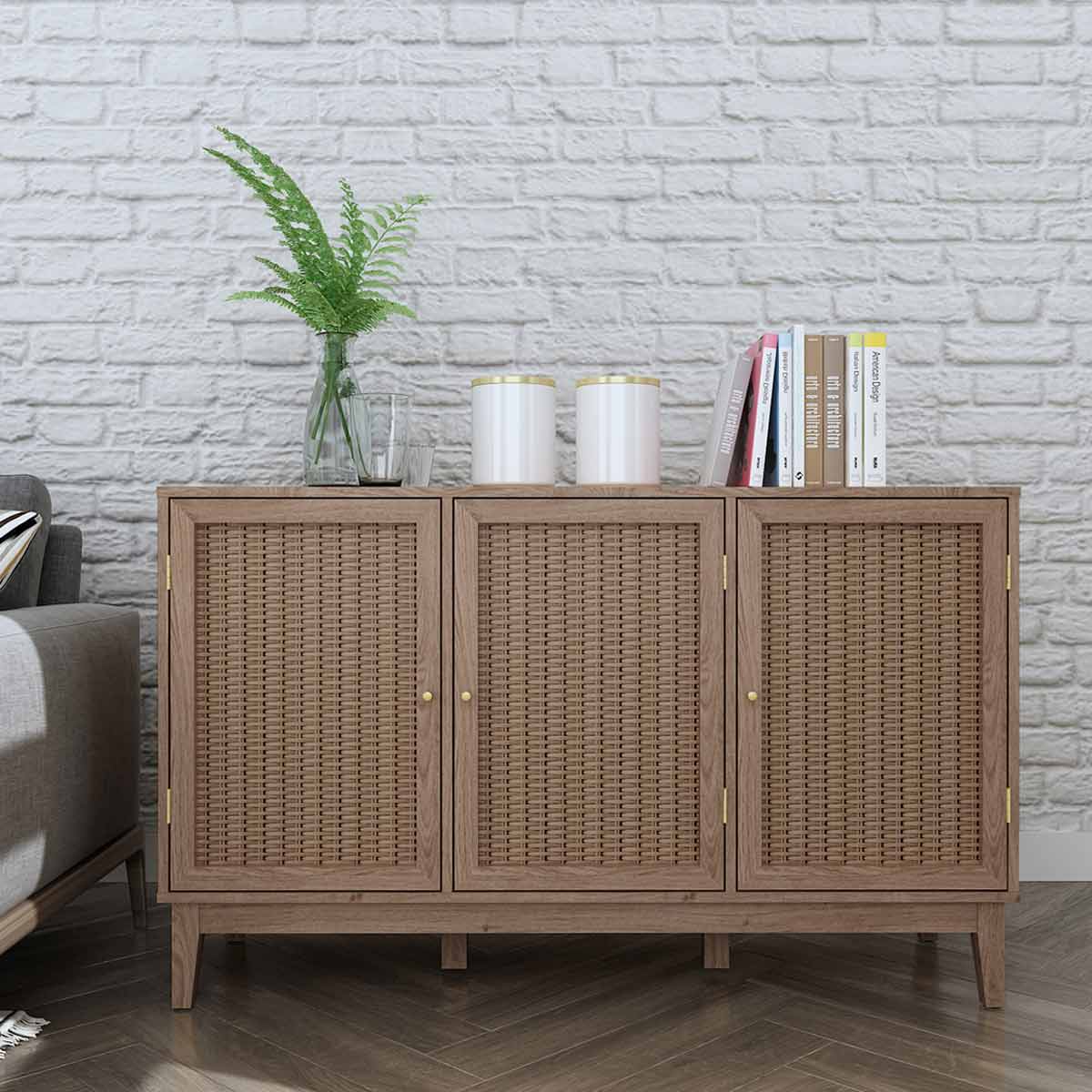 Brown wooden sideboard with three woven rattan doors, gold handles, and a flat top displaying two white canisters and stacked books, set against a textured white brick wall.