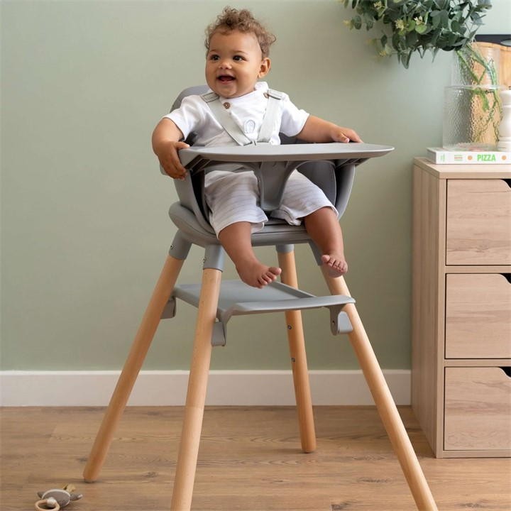 Clair De Lune 6-in-1 high chair in grey with a wooden base, featuring a removable tray and adjustable seat, accommodating a smiling child wearing a white outfit.