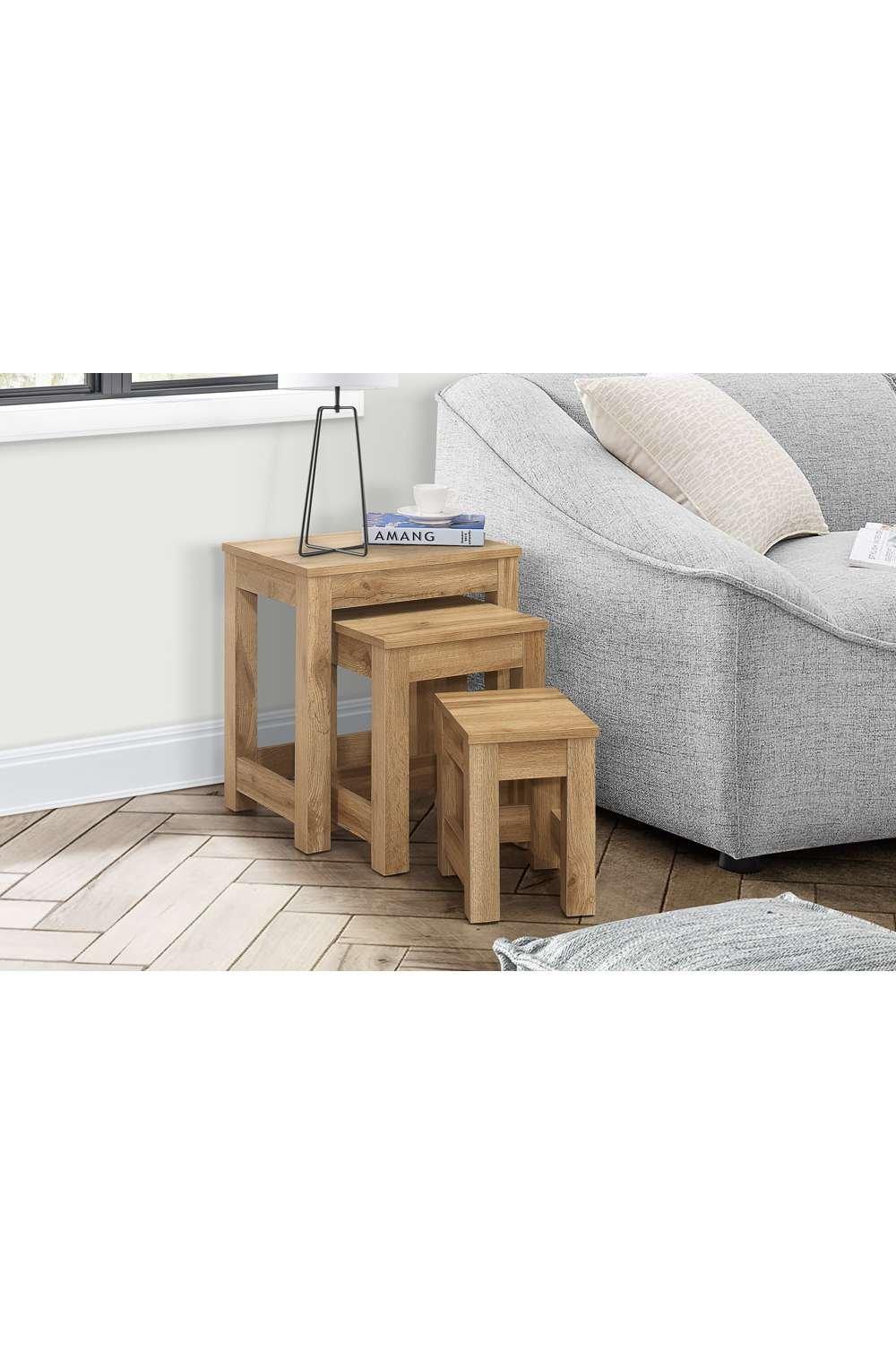 Three brown oak nested side tables with a natural finish, featuring clean lines and a minimalist design, placed beside a light gray sofa and a decorative lamp.