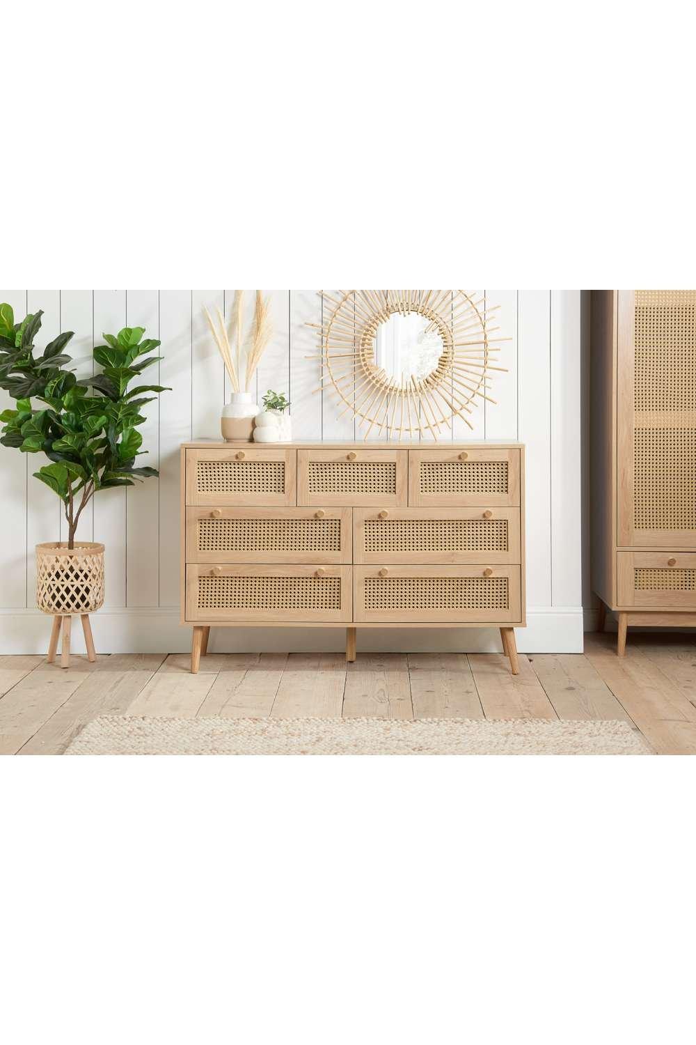 Croxley chest of seven drawers in brown rattan, featuring a mid-century modern design with natural wood legs, complemented by decorative plants and a round mirror.