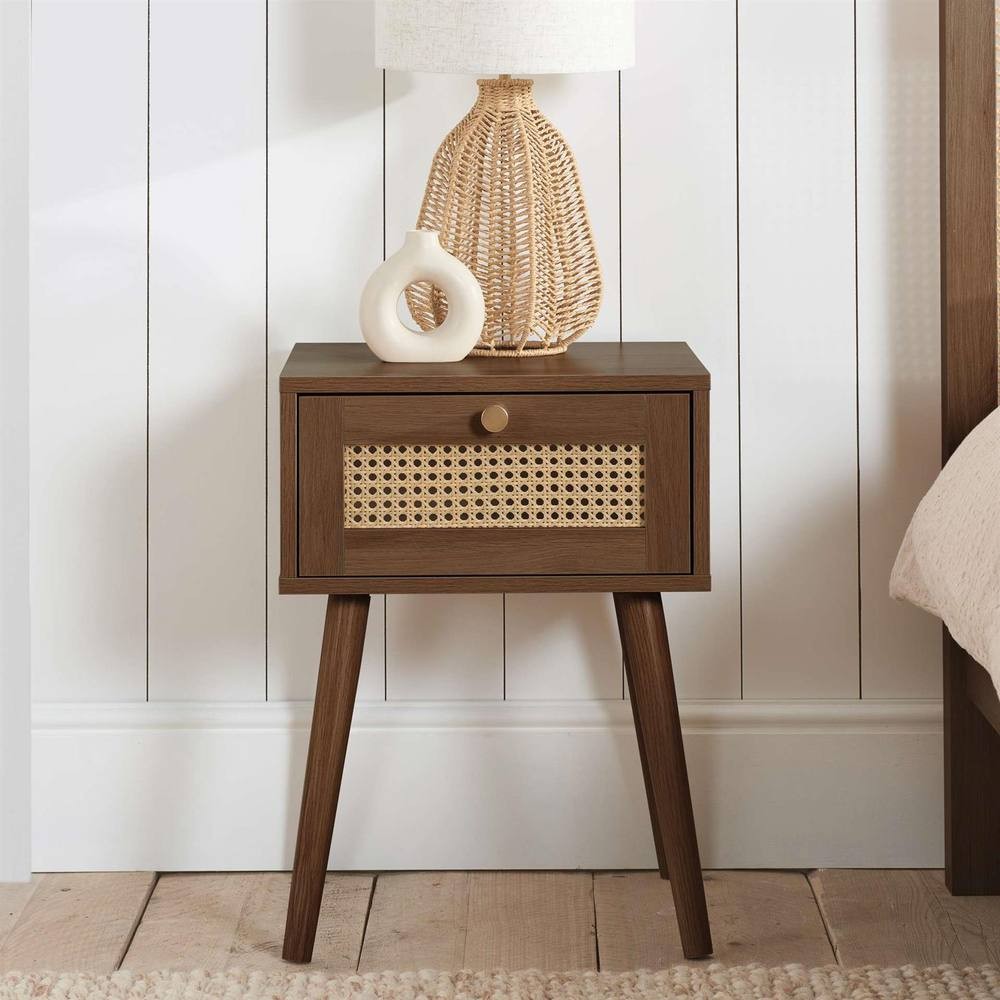 Walnut bedside cabinet with one drawer featuring rattan detailing, supported by tapered legs, accompanied by a textured lamp and decorative vase on top.