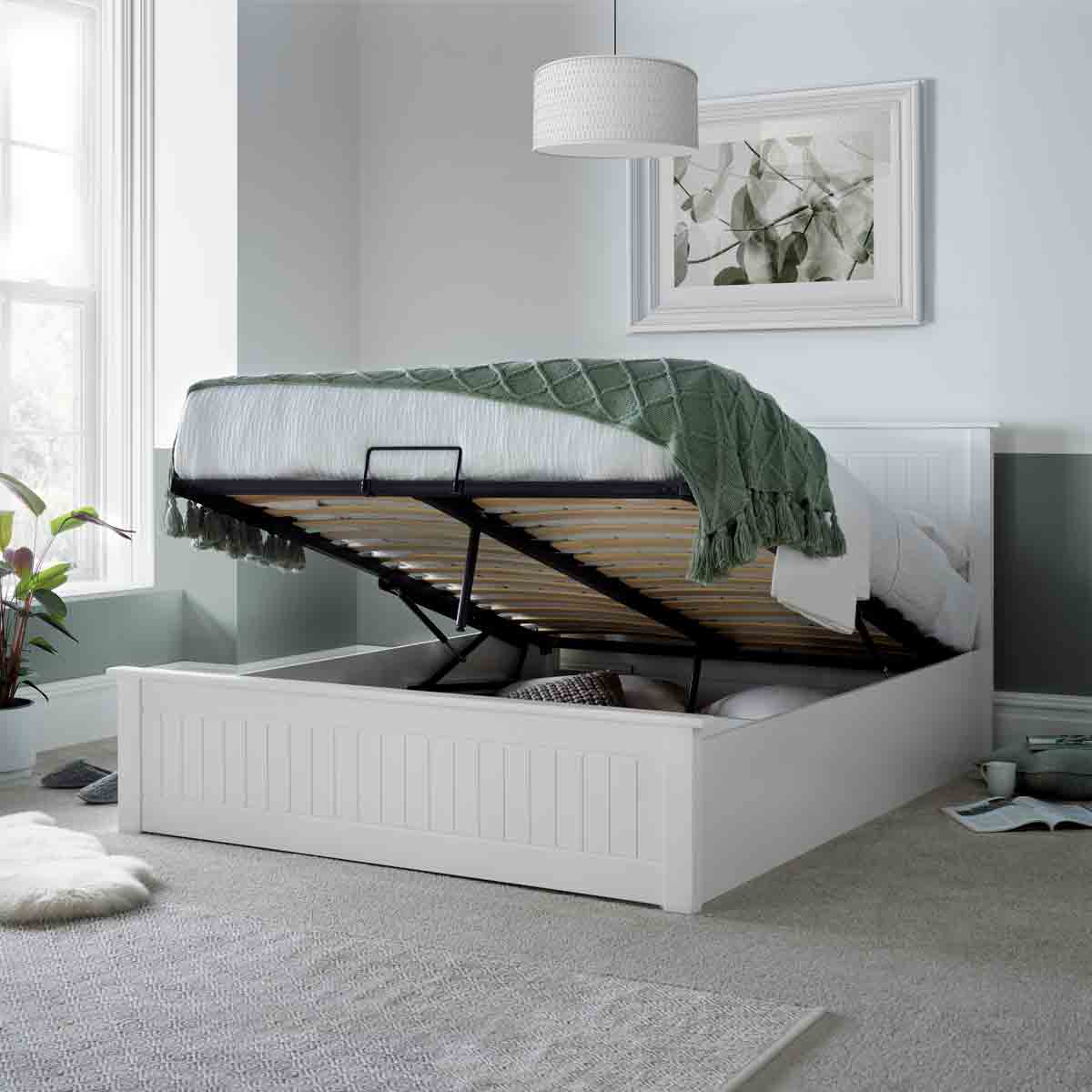 White wooden ottoman bed with a double size, featuring a lift-up mattress revealing storage space underneath, set in a softly lit room with a green throw and decorative plant.