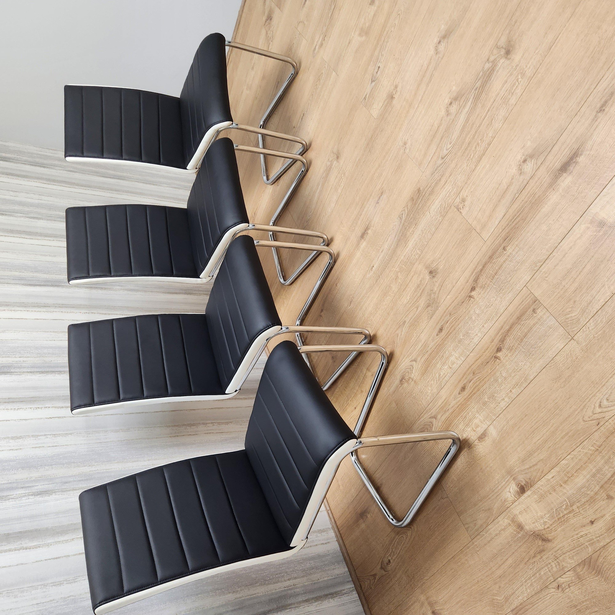 Set of four high-back dining chairs featuring padded faux leather seats in black, with sleek chrome legs, arranged on a light wooden floor.