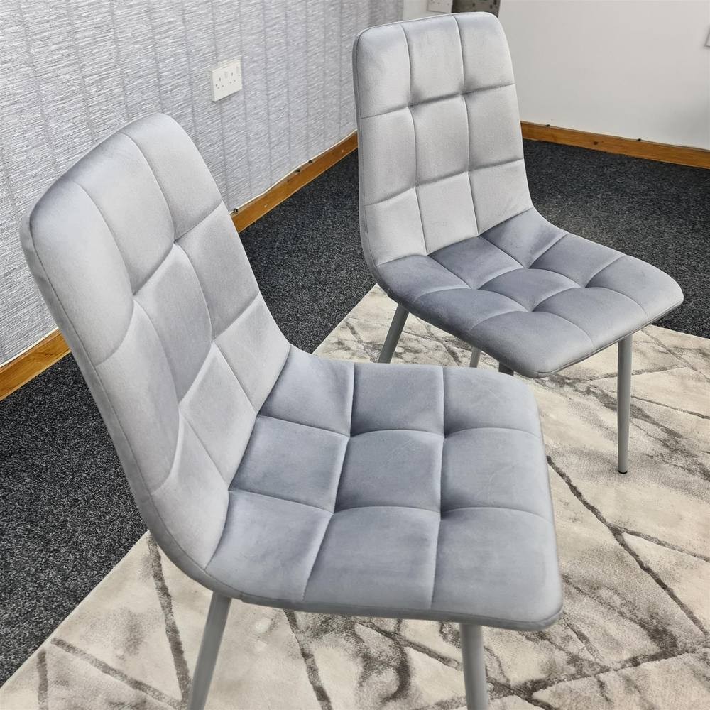 Set of two grey velvet dining chairs featuring a tufted design and sleek metal legs, placed on a patterned rug in a modern interior setting.