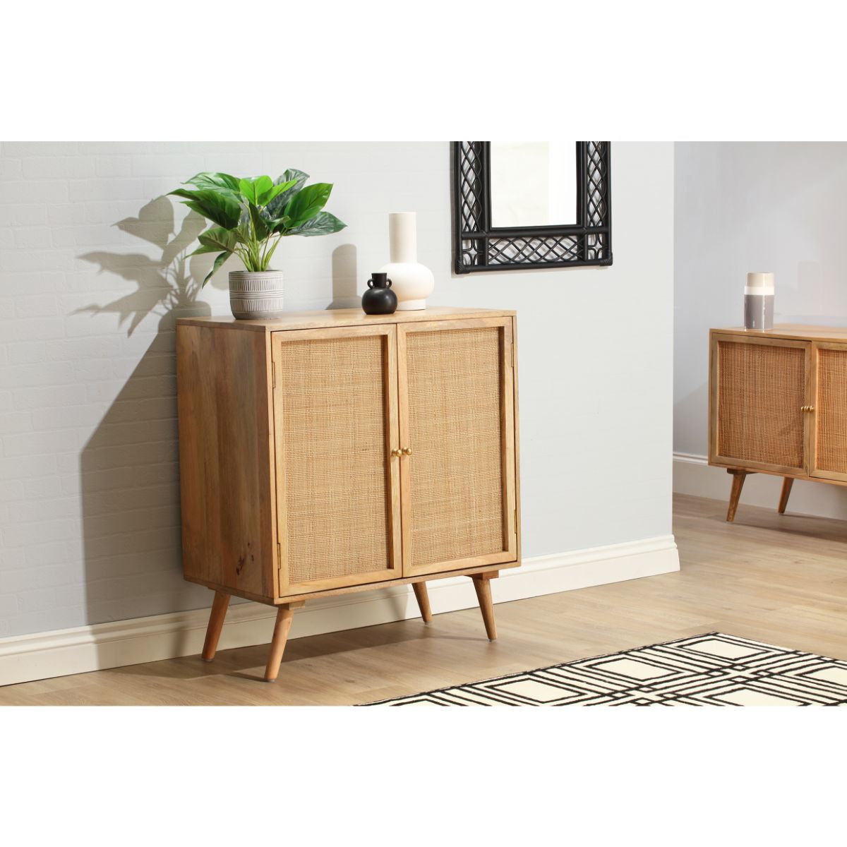 Natural brown rattan sideboard with two doors, wooden legs, and a sleek design, complemented by a potted plant and decorative items on a light-colored wall.