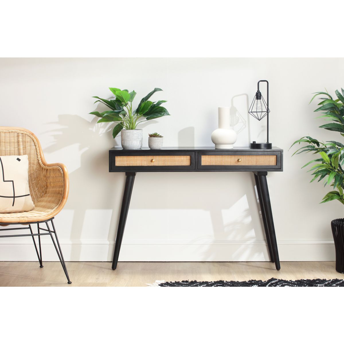 Black console table with two cane drawers, sleek legs, and decorative items like a plant, lamp, and vase, set against a light wall and accompanied by a rattan chair.