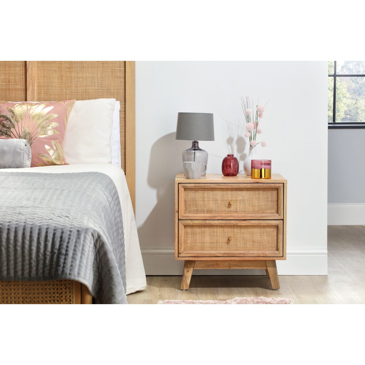 Natural rattan bedside table with two drawers, featuring a light wood finish, styled with a gray lamp, decorative vases, and candles, next to a bed with a gray quilt.