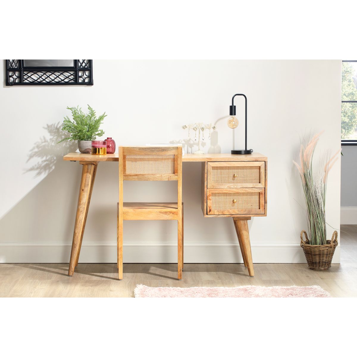 Natural wood desk with two cane drawers and a matching chair, featuring tapered legs, a minimalist design, and a stylish lamp, set against a light wall with decorative plants.
