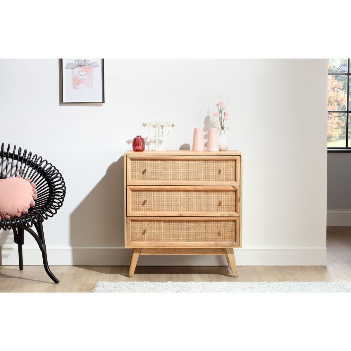 Natural brown rattan chest unit with three drawers, featuring a sleek design and tapered legs, displayed beside a black woven chair and decorative items on a light-colored floor.