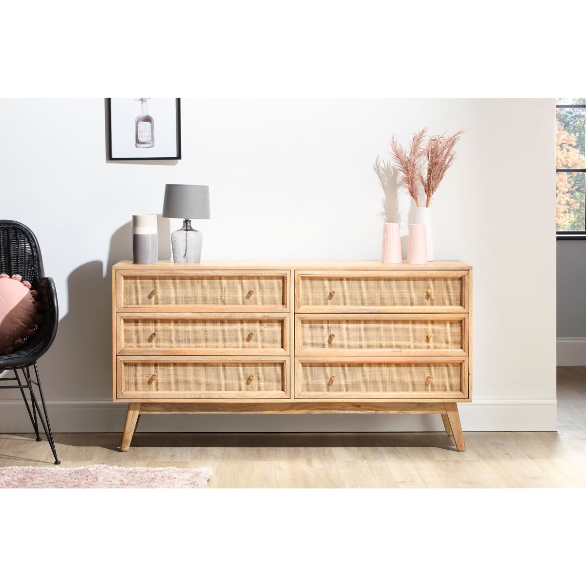Natural brown rattan chest unit with six drawers, featuring wooden handles, set on tapered legs, accompanied by decorative vases and a lamp against a light wall.