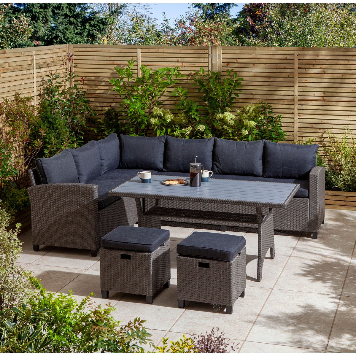 8-seater outdoor dining set featuring a grey wicker finish, plush dark cushions, a rectangular table, and two matching stools, set against a lush garden backdrop.