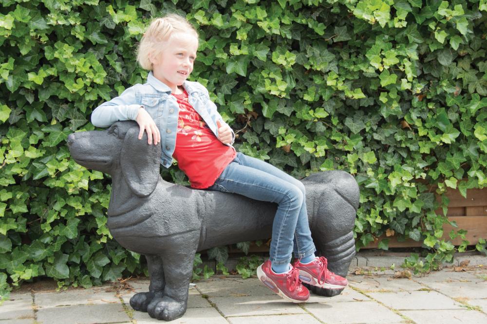 Black fiberstone dachshund bench designed for outdoor seating, featuring a playful dog shape, with a child sitting on it against a backdrop of green ivy.